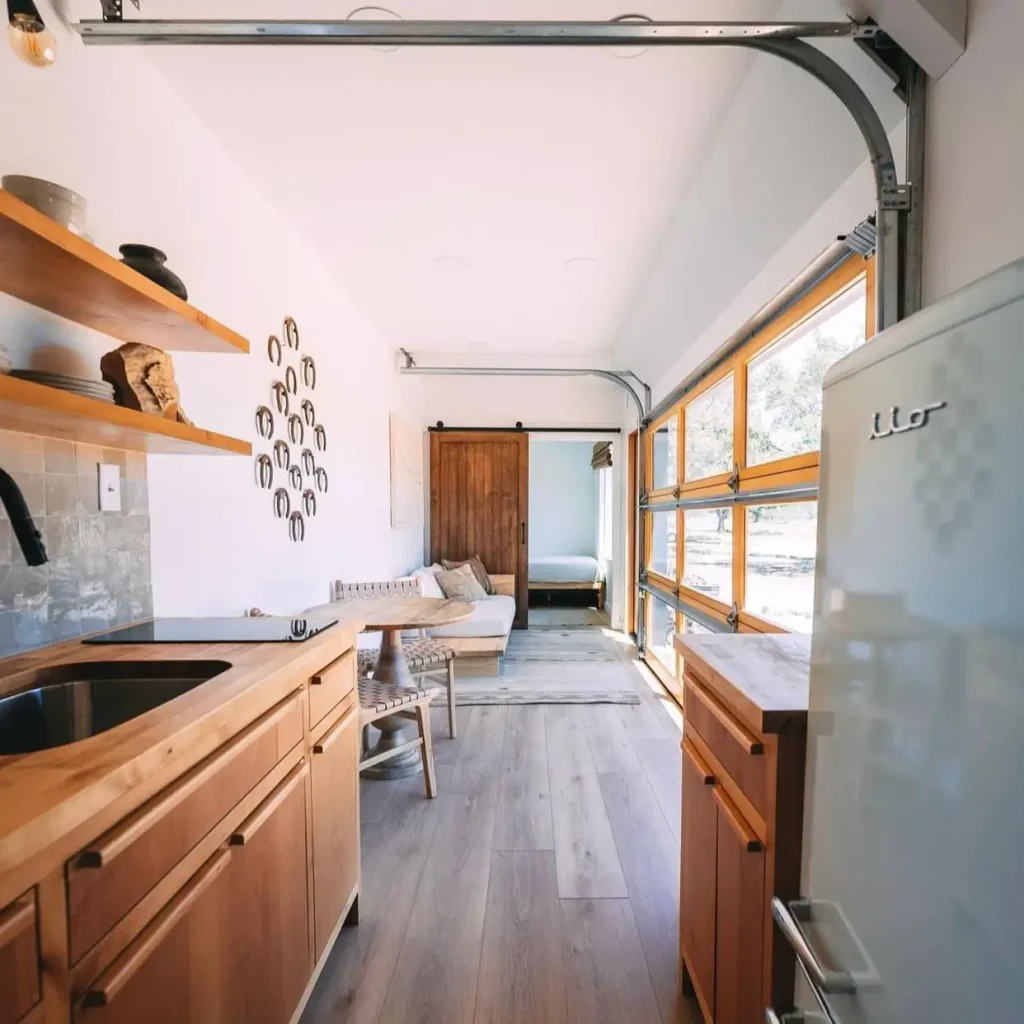 A bright small container house interior featuring a cohesive design with light wood cabinetry, flooring, and a large glass-paneled door.