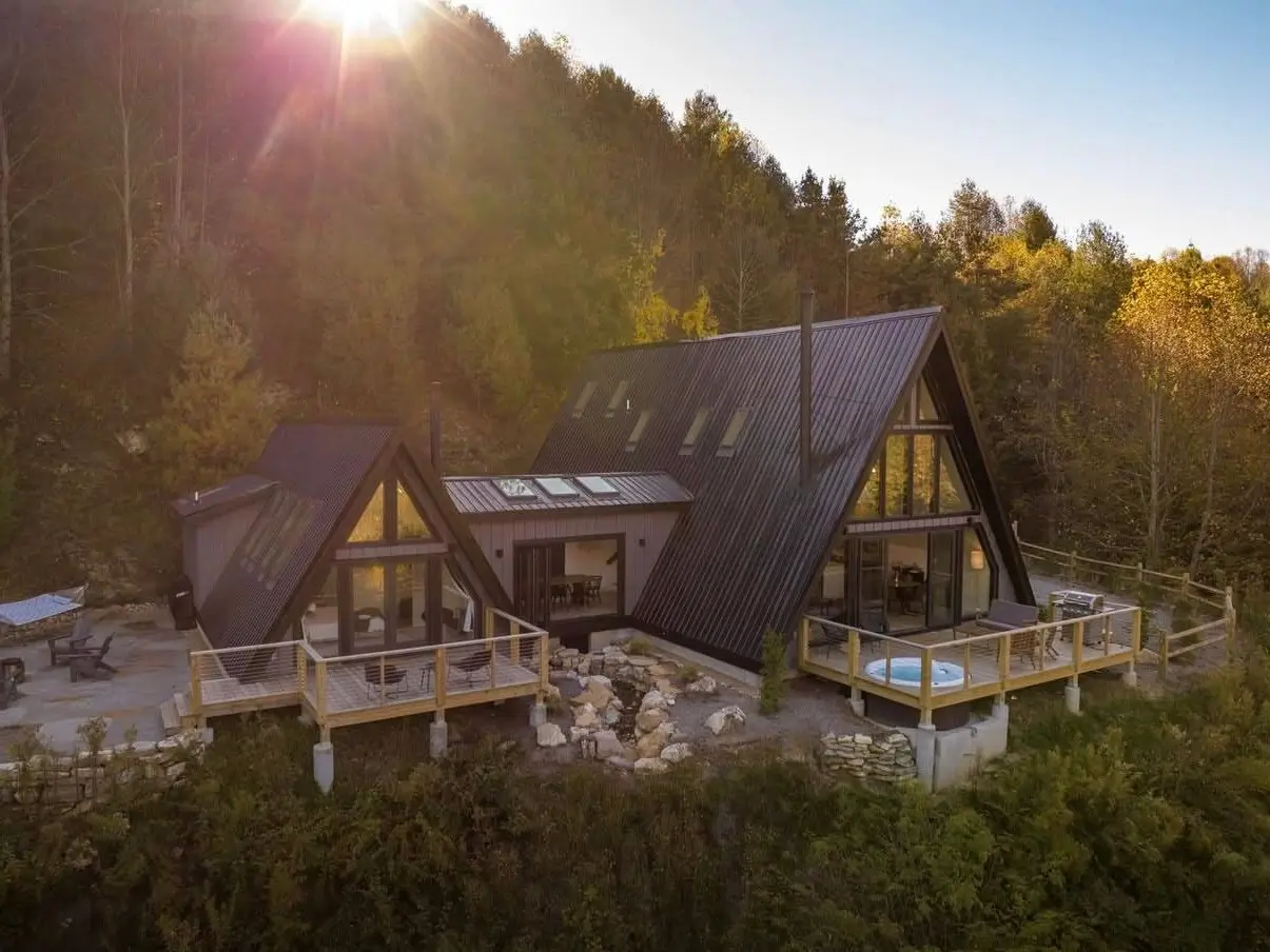 A stunning modern A-frame house compound with multiple peaks and expansive decks with a hot tub, nestled on a sunlit hillside.