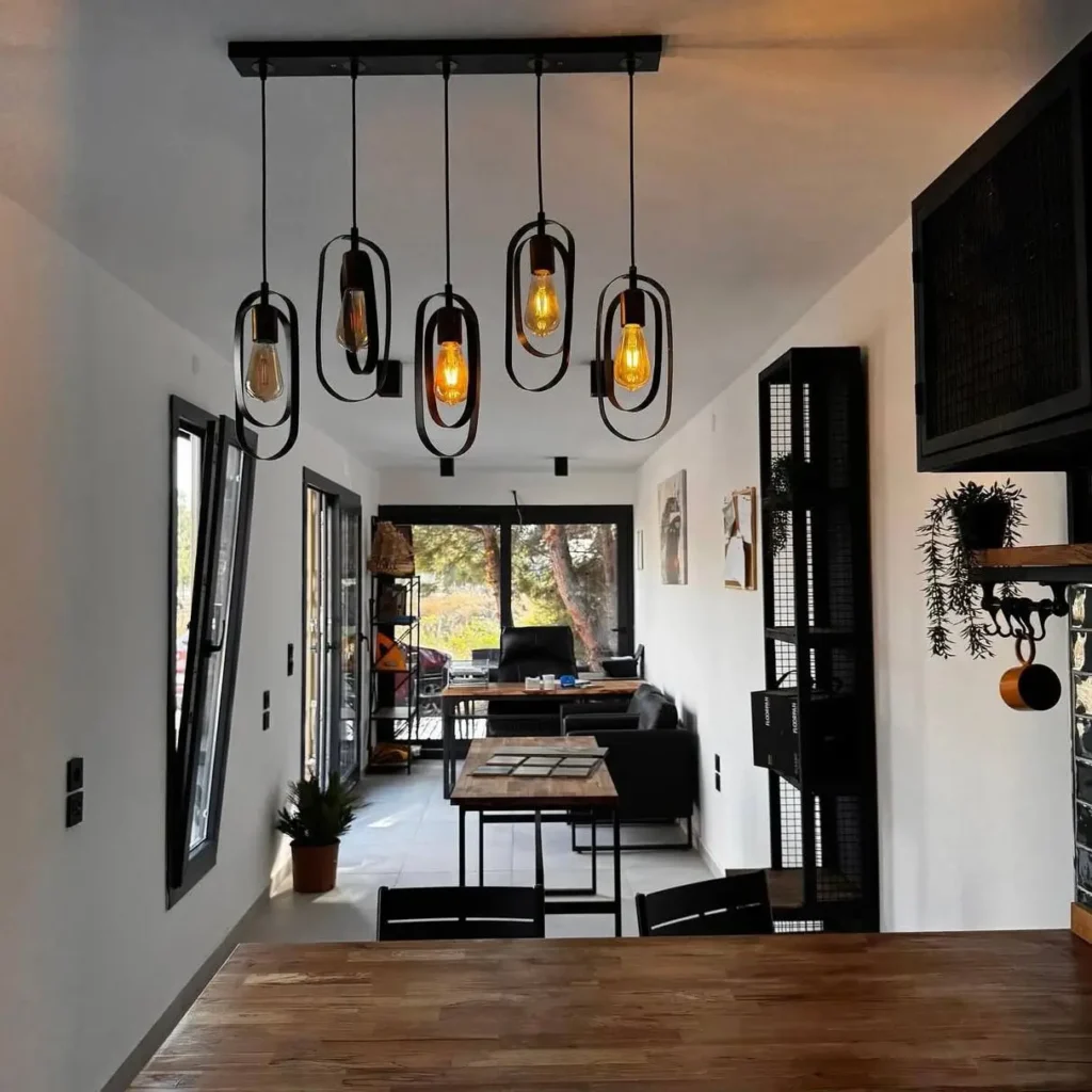 A long, modern-industrial interior of a small shipping container home unified by a dramatic black pendant light fixture over the living and dining areas.