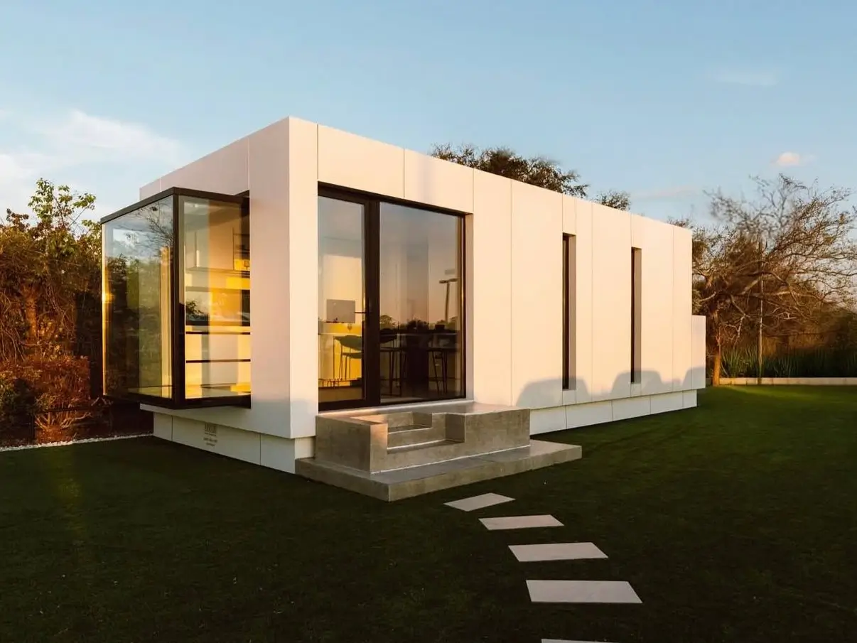 A modern, minimalist tiny house with a white exterior and a large glass corner window, showcasing a unique tiny home design idea.