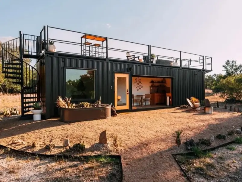 A modern black small shipping container home with a spiral staircase leading to a rooftop deck in a sunny, rustic setting.