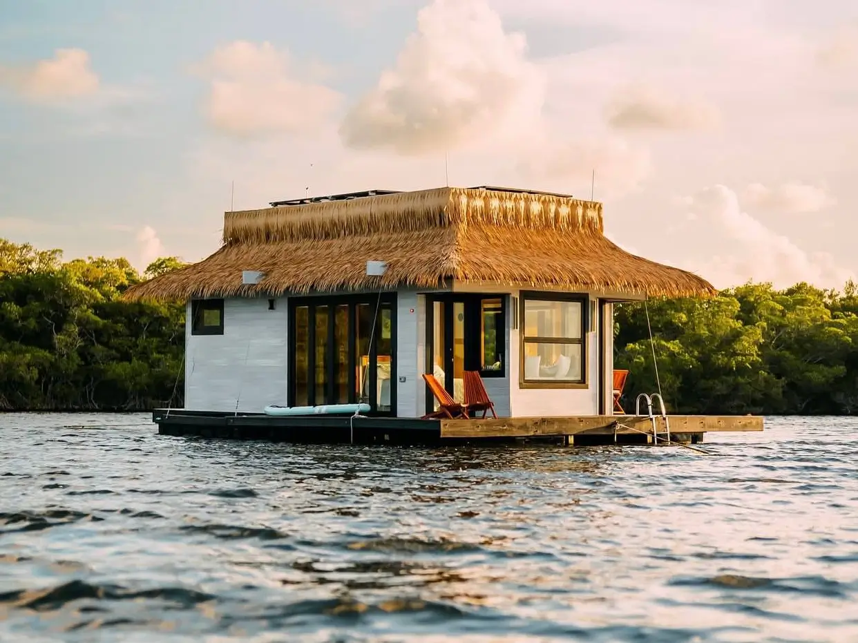 A stunning house design featuring a modern floating home with a thatched roof, sitting peacefully on the water at sunset.