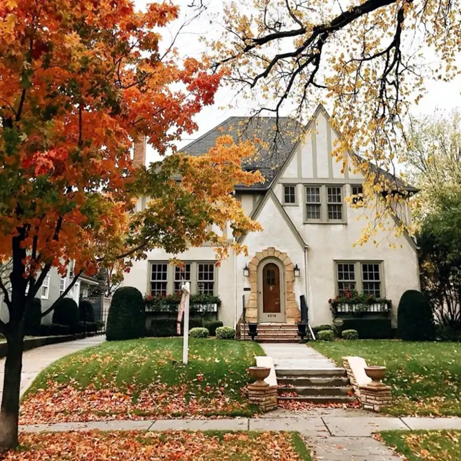A Tudor-style house exterior with a stately stone archway and cream-colored stucco, framed by autumn trees.