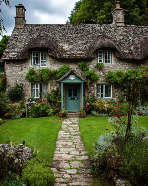 Classic english cottage house design featuring rustic natural stone walls and a traditional thatched roof, with a stone path leading through a lush green garden.