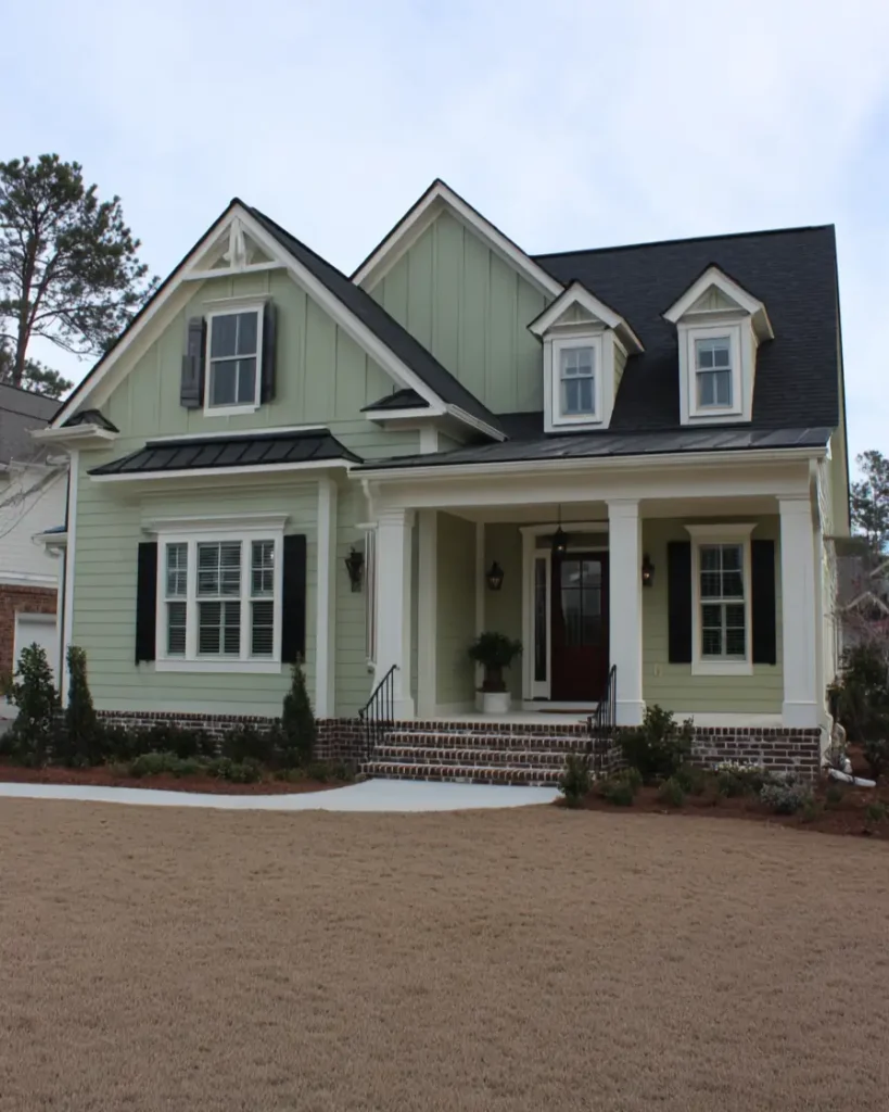 A modern sage green house with black trim, featuring a mix of vertical and lap siding and a welcoming porch with white columns.