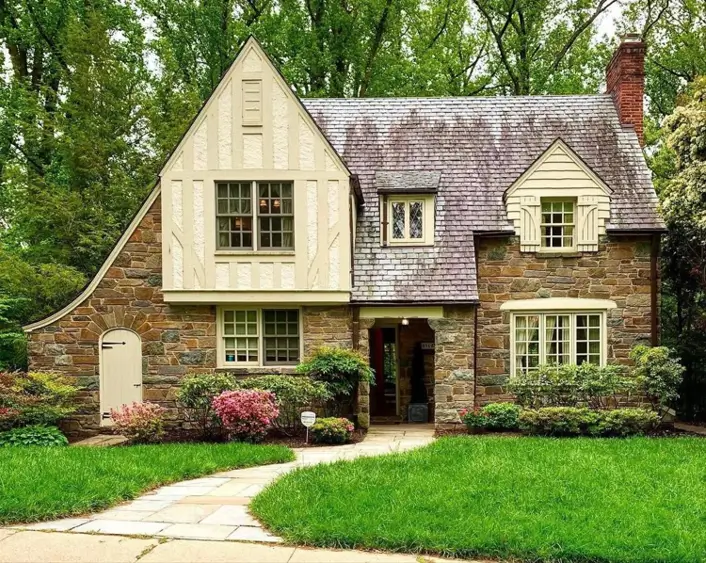 A rustic small Tudor home with a complete fieldstone exterior, a dramatic sweeping roofline, and simple cream-colored gables for a cottage feel.