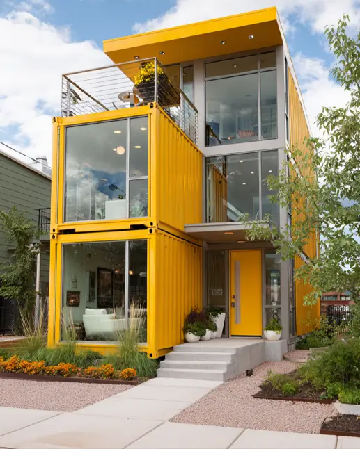 A modern three-story shipping container home painted a vibrant yellow, with large glass windows and a second-floor balcony, showcasing a bold and cheerful design.
