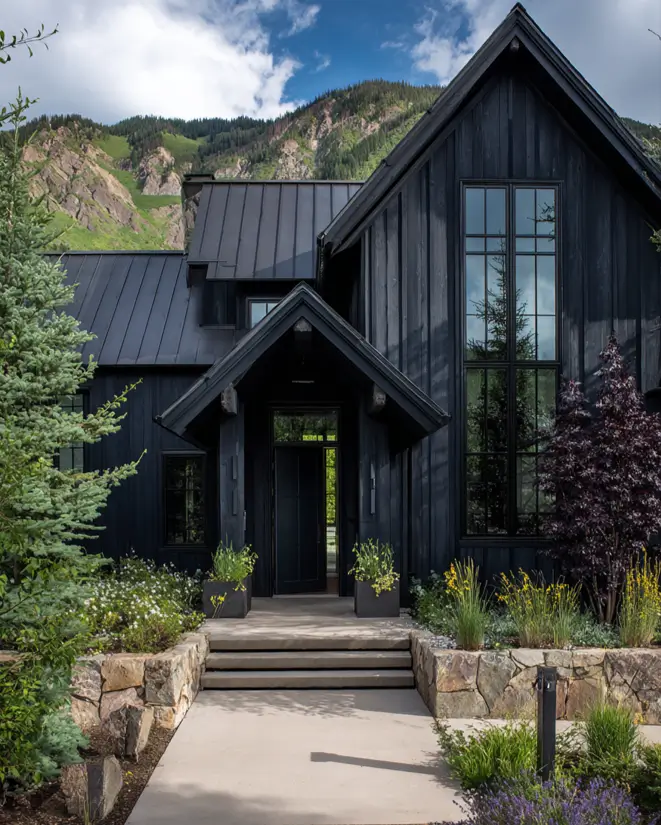 A modern farmhouse painted entirely in black, with a welcoming entryway and lush landscaping against a mountain backdrop.