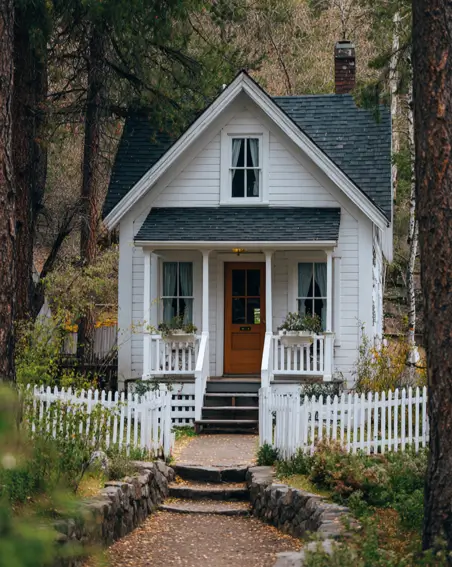 36 Tiny House Design Ideas + An Expert Interior Guide for 2025 17 A classic white cottage with a wooden door and a white picket fence in a forest.