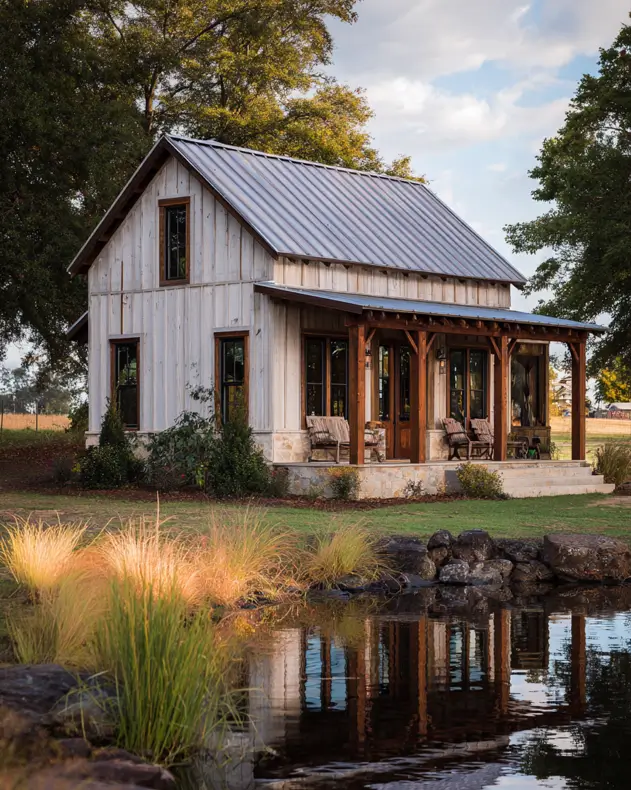 A rustic whitewashed barndominium home with a large wooden porch, situated next to a tranquil pond reflecting the structure.