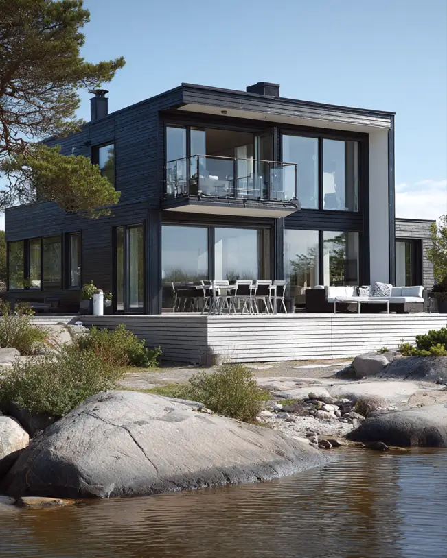 A two-story Scandinavian style home exterior with black siding and a large deck, situated on a rocky shoreline.