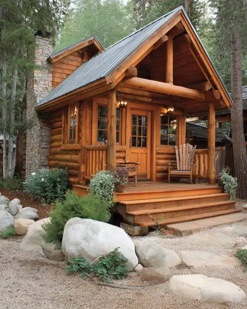 A welcoming tiny log cabin featuring wide entry steps, a stone chimney, and a metal roof, set in a landscaped gravel yard.