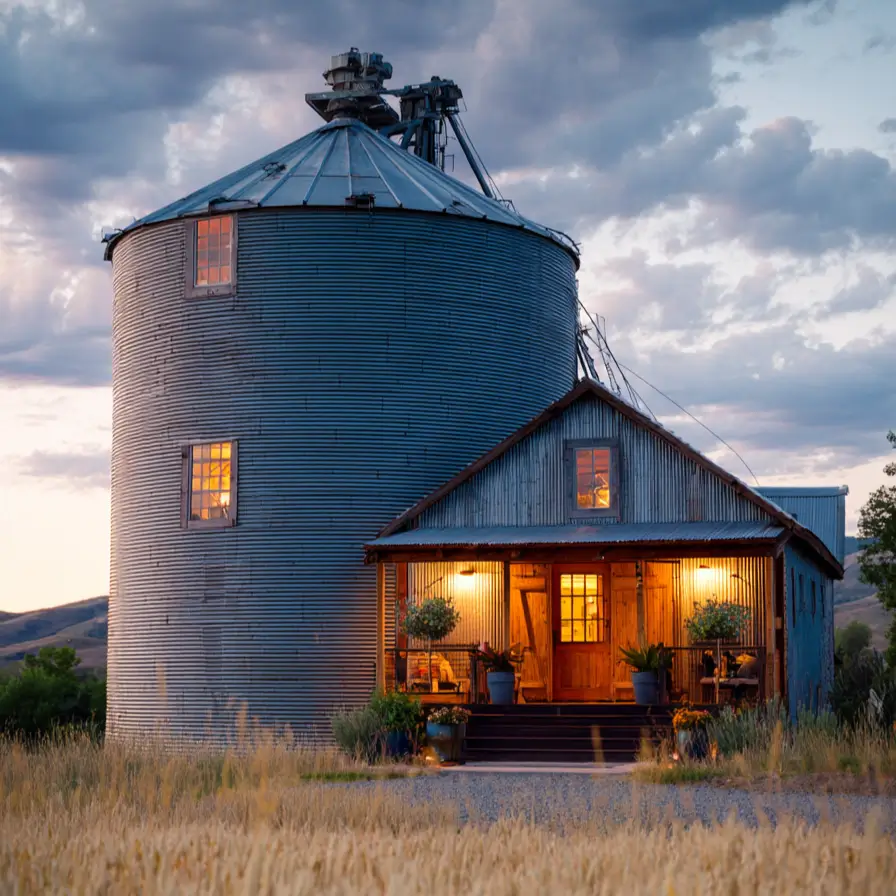 A rustic grain silo house with original machinery on top, attached to a warmly lit wooden barn structure at dusk in a wheat field.