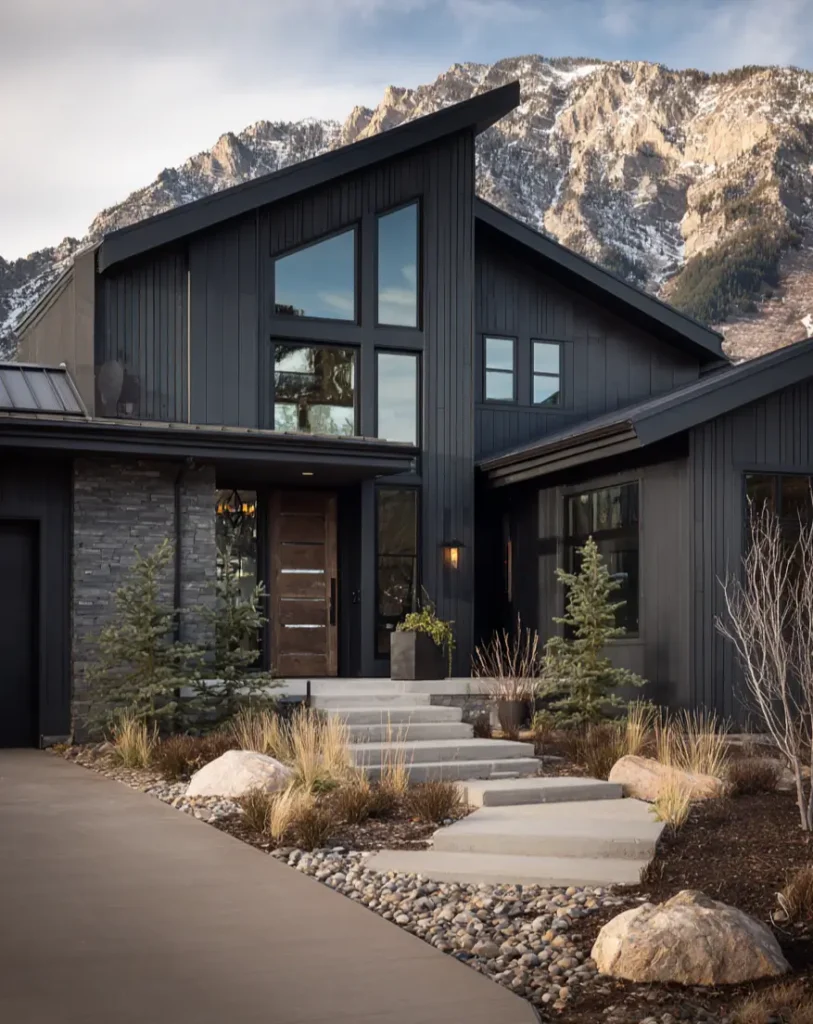 A modern dark gray house with an angled roof and a prominent wooden front door, set against a rugged mountain peak.