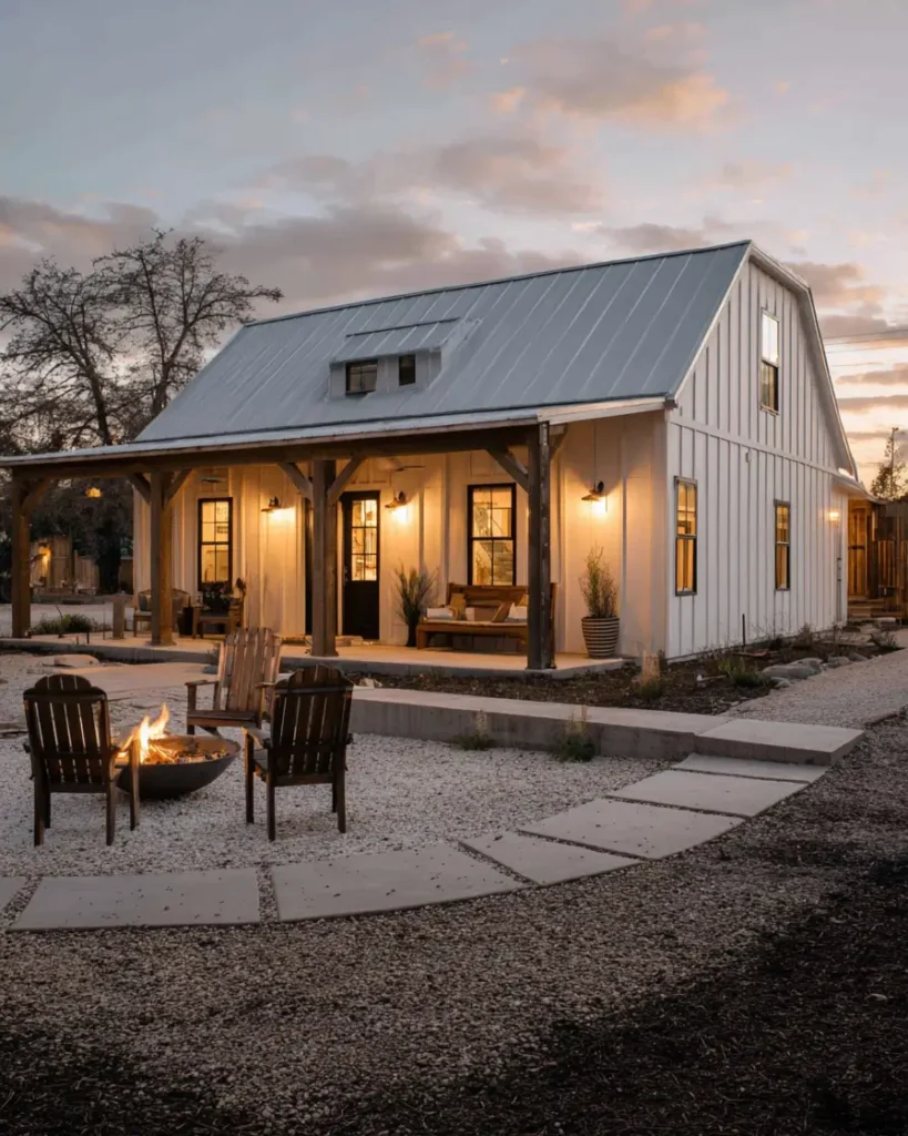 A white barndominium home with a wooden porch, illuminated at dusk, featuring a gravel patio with a fire pit in the foreground.