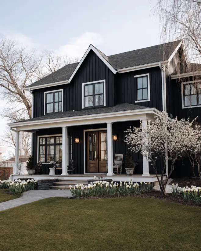 A classic black farmhouse exterior with a full-width white porch, contrasted by blooming spring flowers.