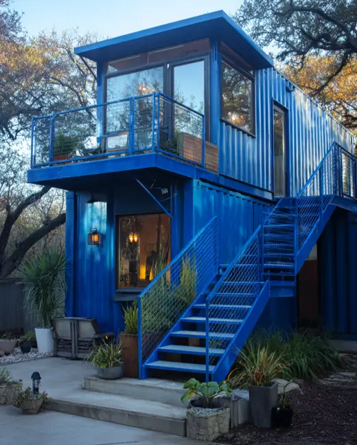 A two-story shipping container home painted a vibrant blue, featuring a matching blue exterior staircase leading to a second-floor balcony, set in a backyard garden setting.