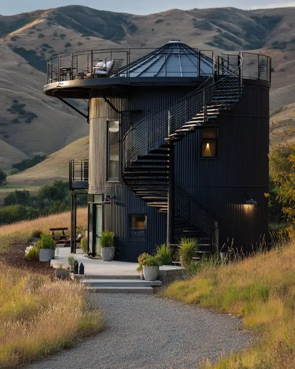 A modern, black grain silo cabin with an external spiral staircase leading to a rooftop deck overlooking rolling hills.