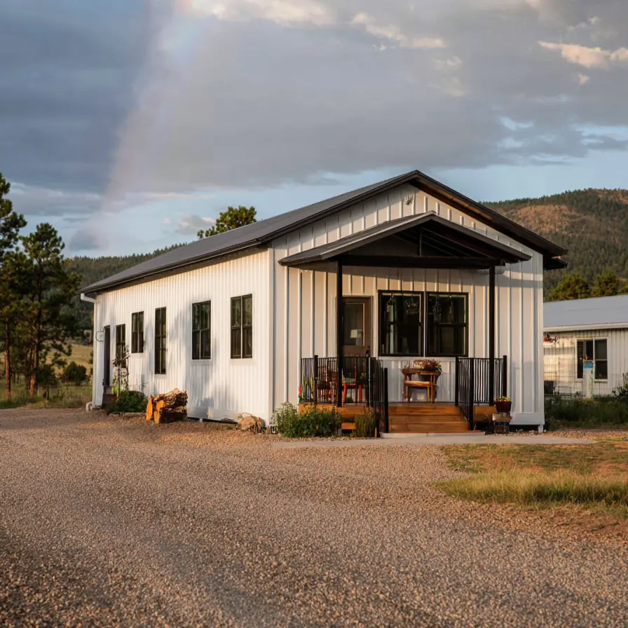 A simple white barndominium home with a black porch and roof, with a bright rainbow in the sky above.
