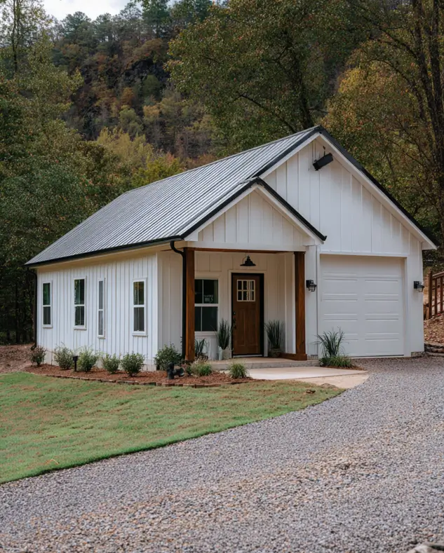 A small white barndominium home with an attached single-car garage and wooden porch posts on a gravel driveway.