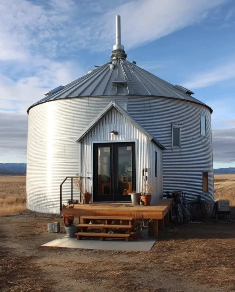A wide grain bin house with a simple gabled entryway and a small wooden deck, set in an open field under a blue sky.
