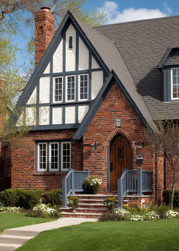 A brick Tudor-style house exterior with sophisticated slate-blue trim, a timbered gable, and an arched wood door.