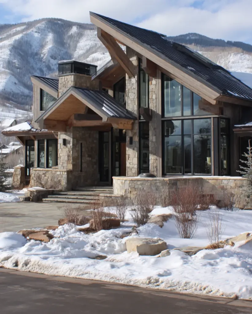 A modern mountain home with a dramatic angled roof, featuring large exposed timber beams and stone walls in a snowy setting.