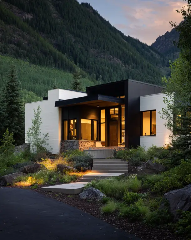 A modern mountain house at dusk with a minimalist white and black exterior, set against a steep forested mountain.