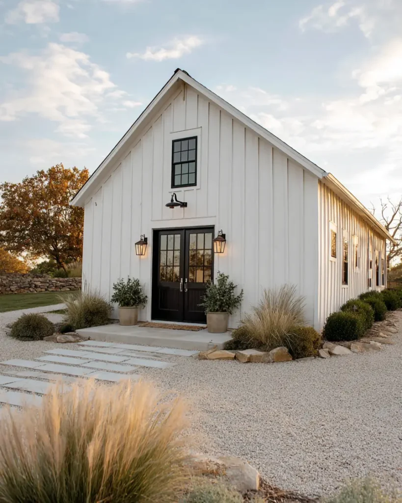 A white barndominium with black double doors, surrounded by a gravel courtyard and a walkway made of stone pavers.