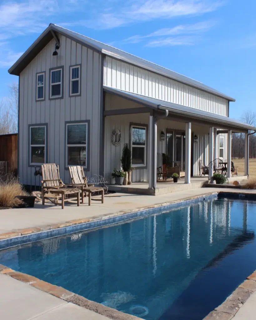 A light gray barndominium home with a long porch overlooking a large in-ground swimming pool on a sunny day.