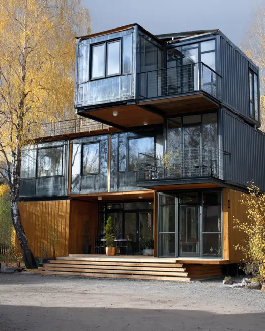 A complex multi-story shipping container home with a dark gray finish and a wooden base, featuring multiple levels of balconies and large glass windows reflecting autumn trees.