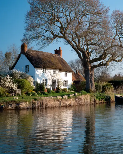 A serene english cottage style home with a thatched roof sitting on the banks of a calm river, its reflection visible in the water.