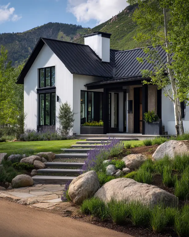 A modern white farmhouse with a black metal roof, surrounded by natural landscaping with boulders and lavender.