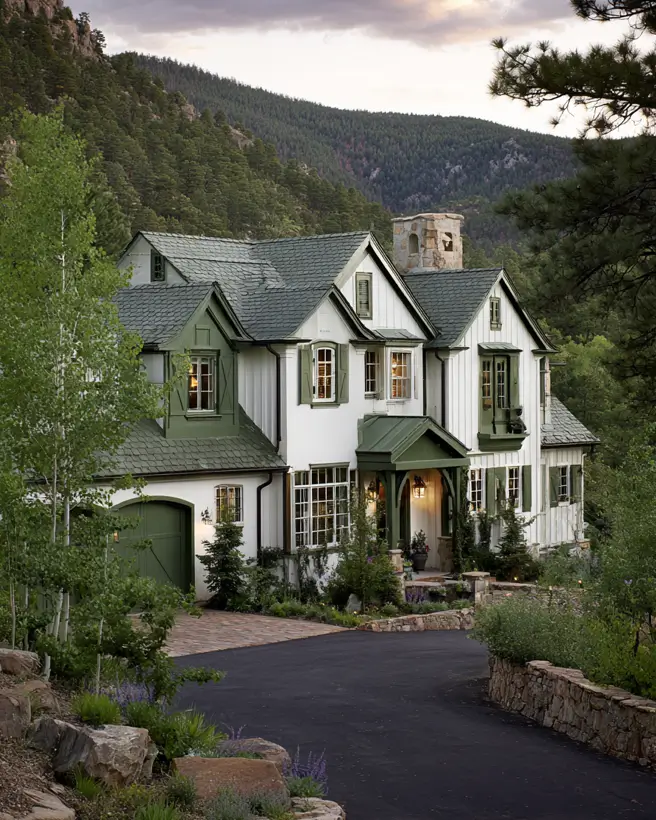 A large white modern mountain house with sage green trim and shutters, surrounded by lush green trees and a hillside.