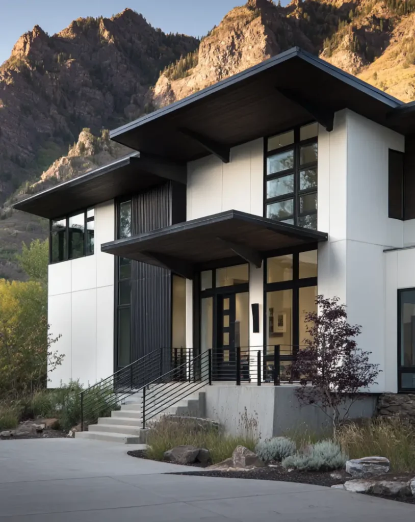 A modern house with a white exterior and dark, overhanging roofs, set against a sunlit, rugged mountain.