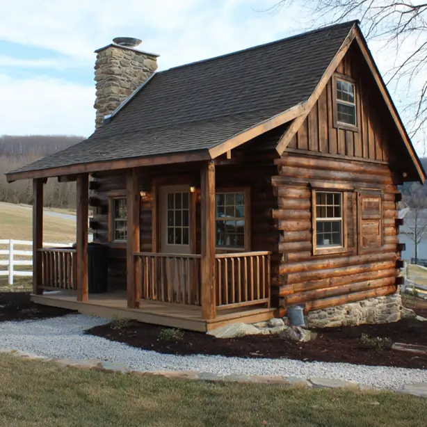 A tidy tiny log cabin home with a wraparound porch and stone chimney, overlooking a rural landscape with a white fence.