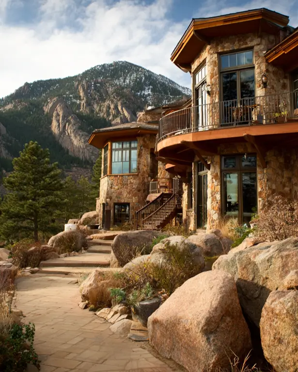 A sprawling stone mountain house with a curved balcony, built into a rocky hillside with a mountain view.