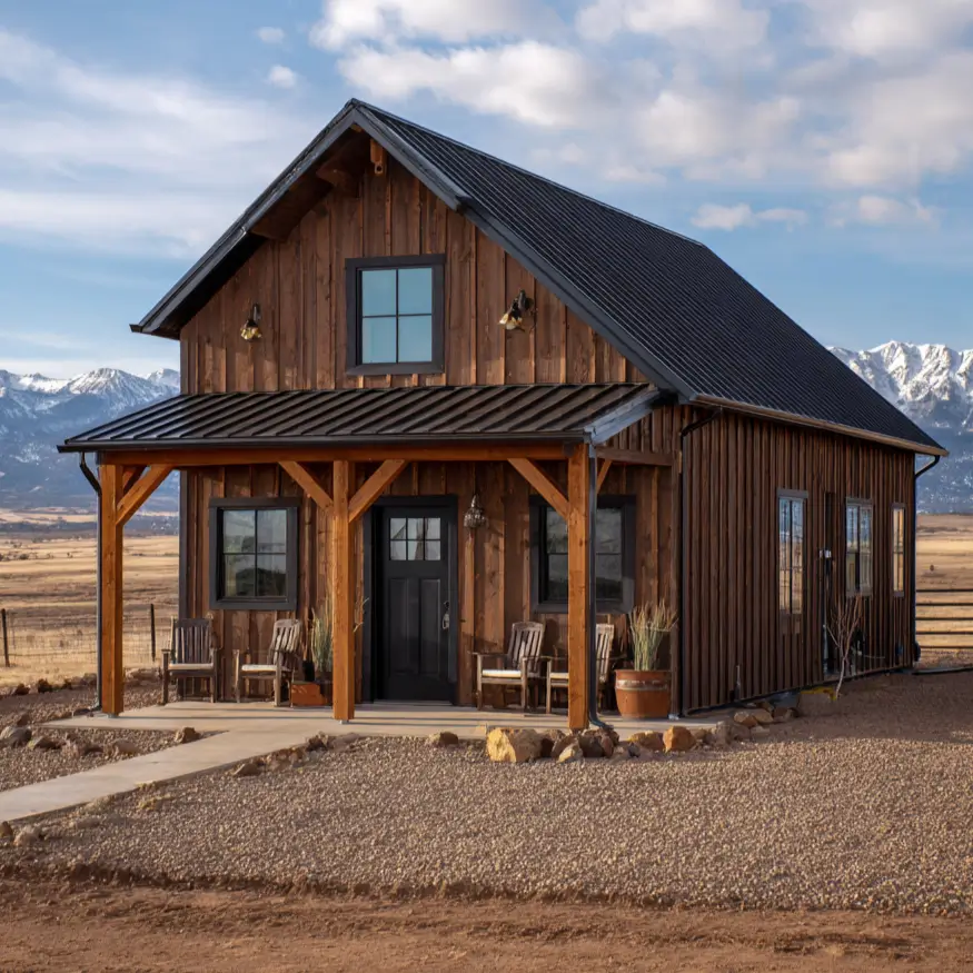 A dark wood barndominium with a prominent timber porch, set in a wide-open landscape with snow-covered mountains in the distance.