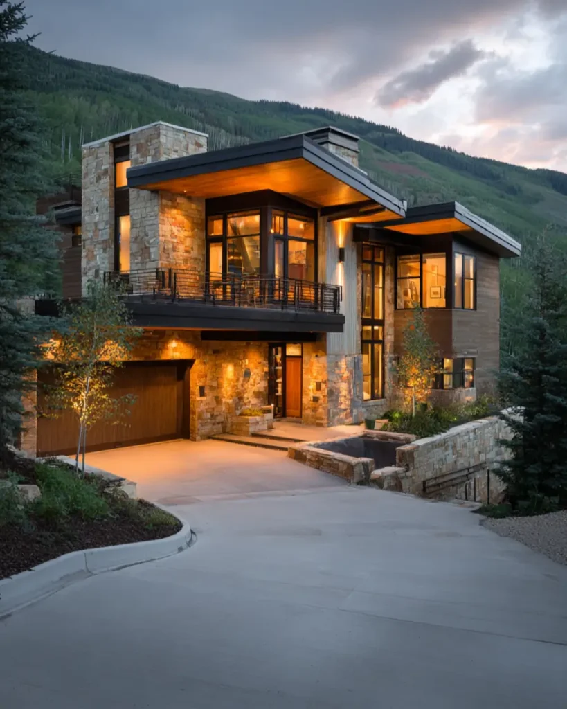 A luxury modern mountain house at dusk, with warm lights illuminating its stone and wood exterior against a forested hillside.