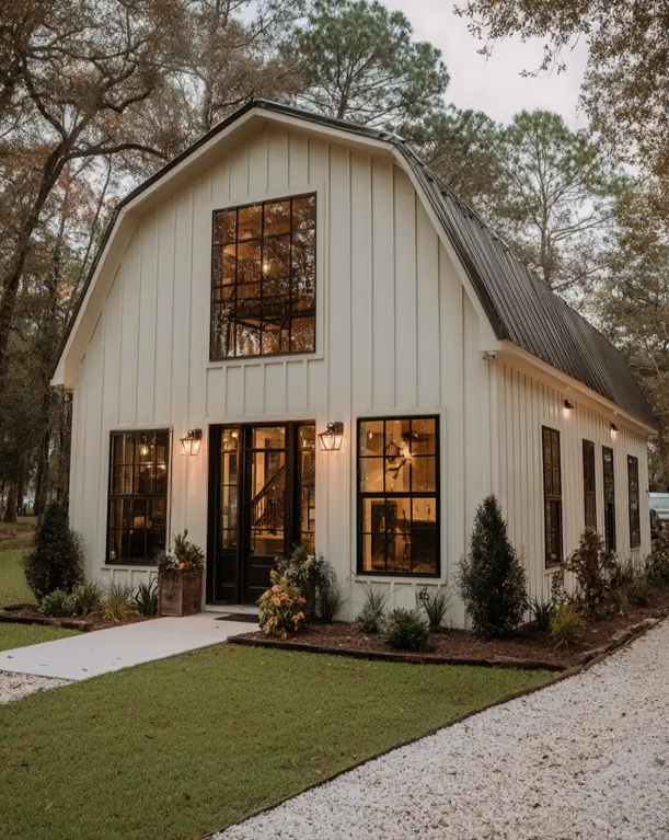 A creamy white small barndominium house with a dark gambrel roof and large, black-framed windows that illuminate the interior at dusk.