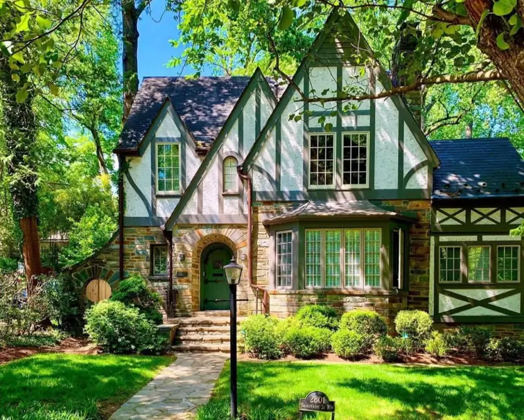 Picturesque small Tudor-style house featuring a rustic stone and stucco exterior with muted green trim that complements the surrounding trees.