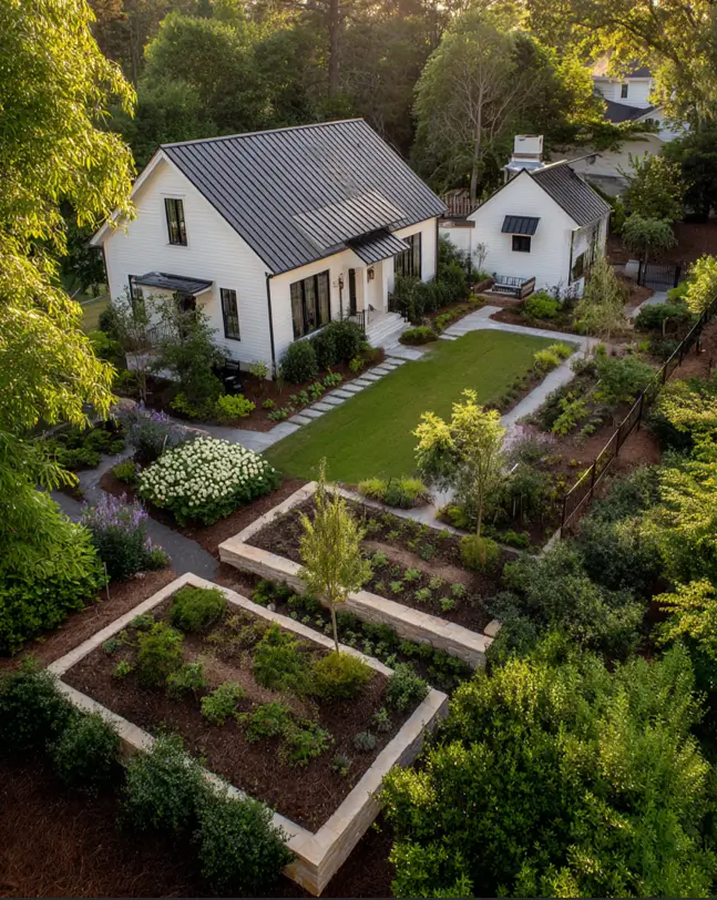 An aerial view of a white and black modern farmhouse connected to a matching guest house by a lush garden.