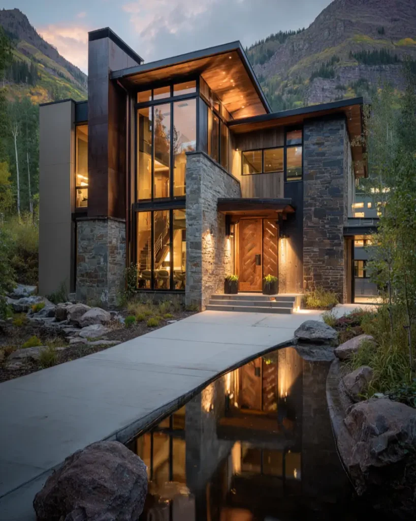 Luxury modern mountain home at dusk with stone and wood exterior, its lights reflecting in a water channel leading to the front door.