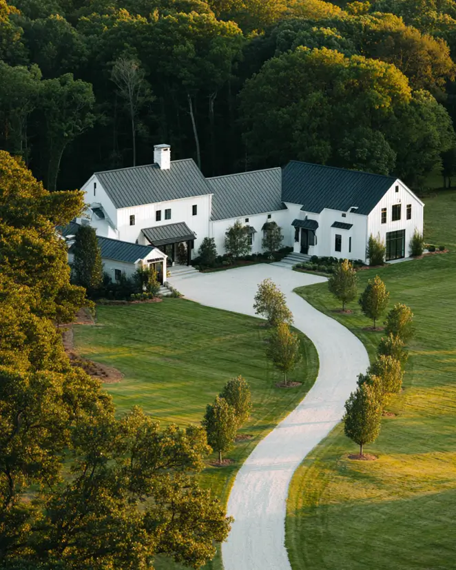 An aerial view of a sprawling white modern farmhouse with a long, winding driveway through a green landscape.