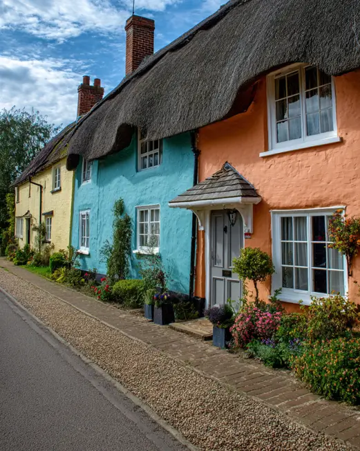 A vibrant row of english cottage style homes with thatched roofs, painted in cheerful shades of yellow, blue, and orange along a village street.