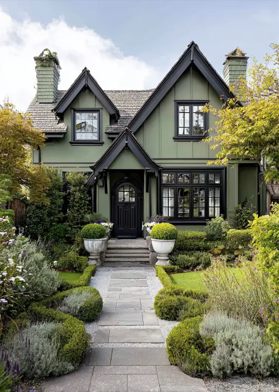 A dark, moody sage green Tudor-style house with dramatic black trim, set within a storybook-like formal front garden.