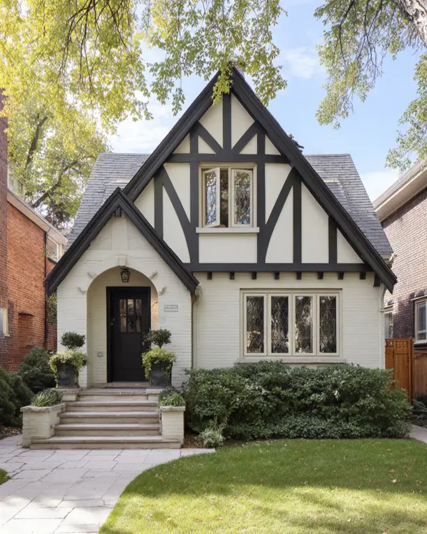 An elegant small Tudor-style house with a refined look, featuring a cream painted brick exterior, simple black timbering, and a light stone arched entryway.