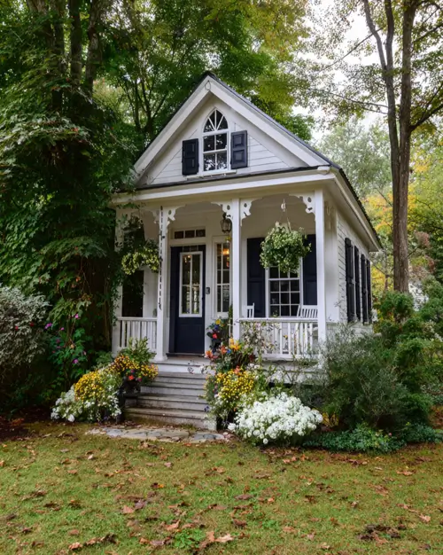 A small white cottage with a covered porch, black shutters, and overflowing hanging baskets and planters.