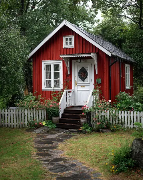 A small red cottage with vertical siding, a white door and picket fence, and decorative trim.