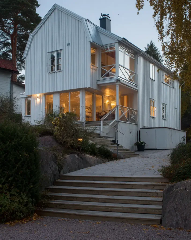 A traditional white Scandinavian house with a gambrel roof, featuring glass-enclosed porches on multiple levels, lit warmly at dusk.